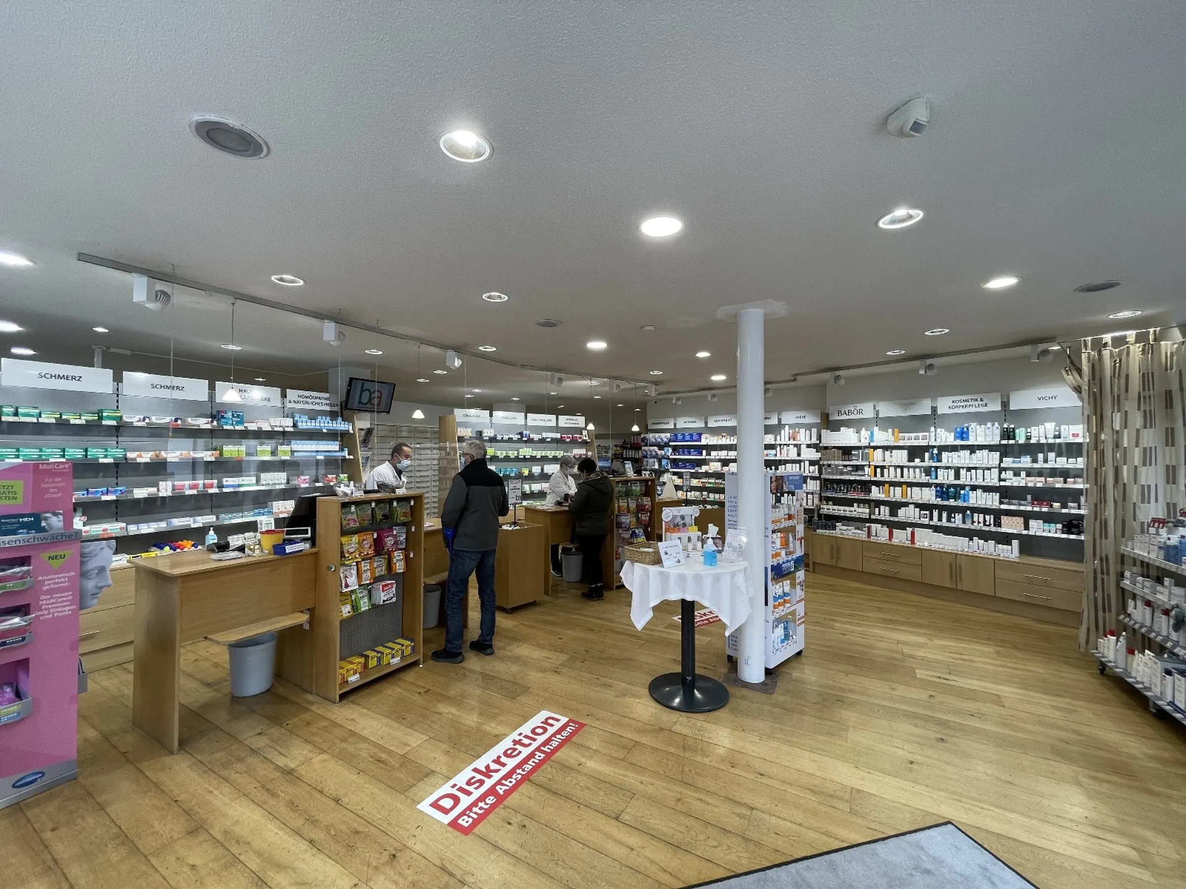 Innenansicht einer Apotheke mit Regalen voller Medikamente, zwei Kunden an der Kasse und einem „Diskretion“ Bodenaufkleber.
