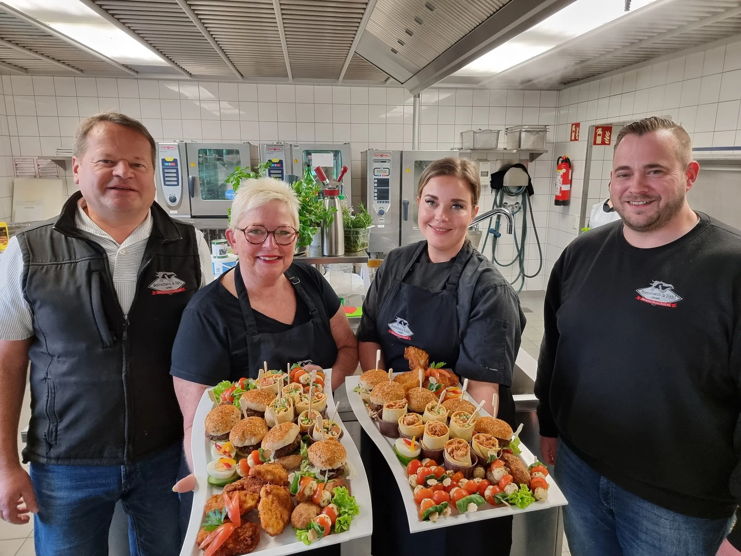 Vier Catering-Mitarbeiter in schwarzer Kleidung präsentieren zwei Platten mit verschiedenen Häppchen in einer Küche.