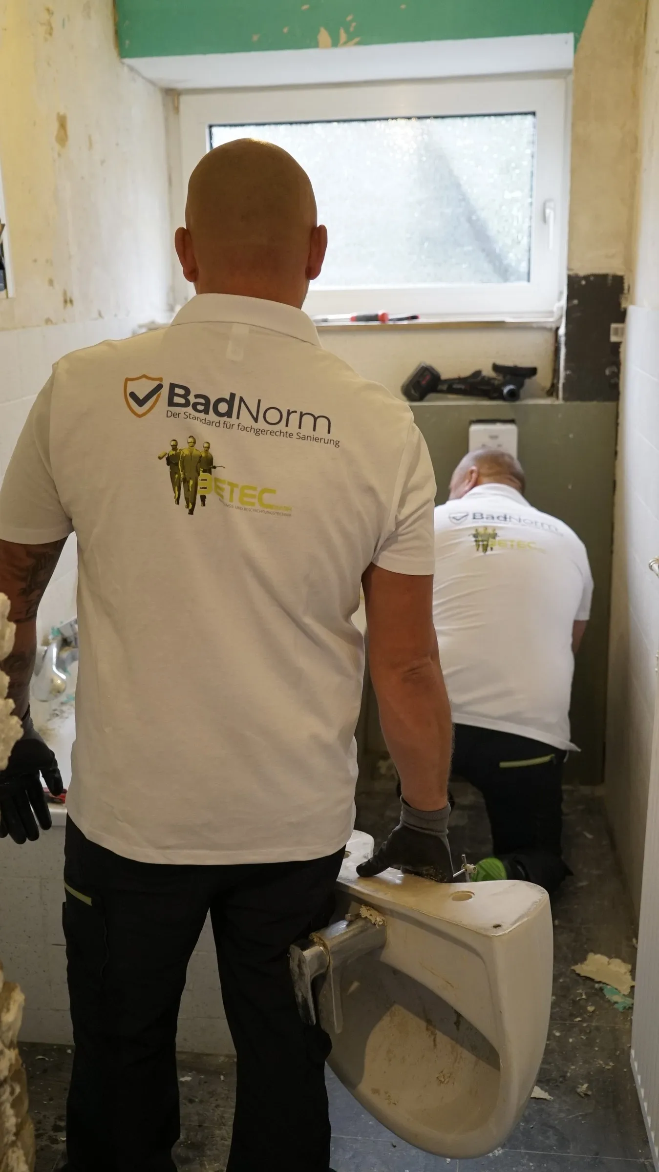 Zwei Handwerker in BadNorm-T-Shirts entfernen eine Toilette aus einem Badezimmer.