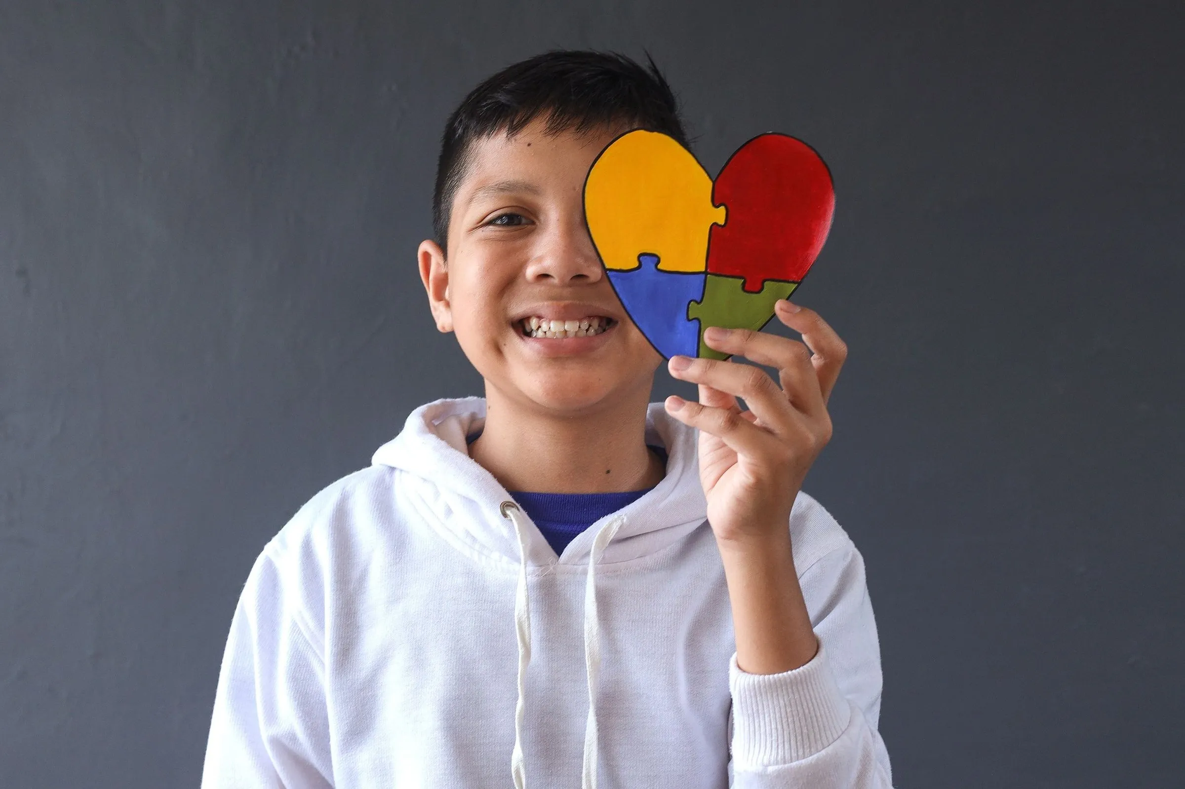 Lächelnder Junge in weißem Hoodie hält ein herzförmiges Puzzle mit vier bunten Teilen vor sein Gesicht.