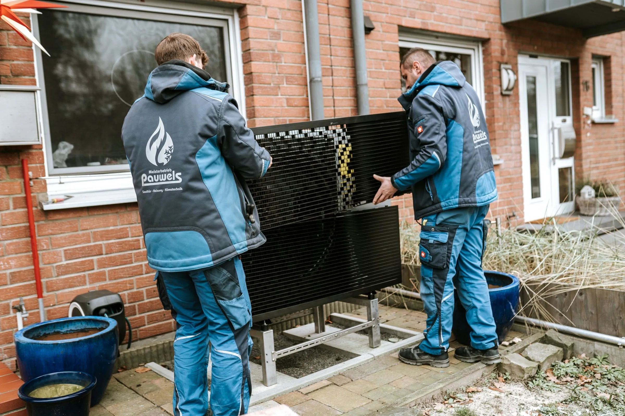 Zwei Installateure in blauen Jacken mit "Pauwels"-Logo montieren schwarze Solarthermie-Kollektoren an einem Haus.